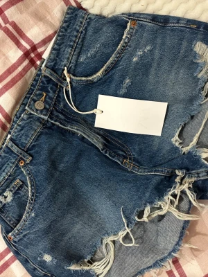 Blå jeansshorts med fransar - Säljer ett par blå denimshorts med slitna detaljer och fransig kant. Shortsen har klassisk femficksmodell, knappgylf och råa avslut. Aldrig använda. 