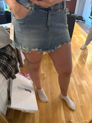 Blå jeanskjol med fransig kant - Säljer en kort blå jeanskjol med fransig och sliten kant. Kjolen har klassisk femficksmodell och råa detaljer längst ner för en trendig look. Aldrig använd, lappen kvar 