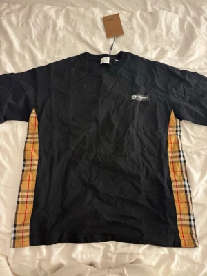 Svart t-shirt med rutiga sidopaneler - Snygg svart t-shirt med klassiska beige-rutiga sidopaneler och korta ärmar. T-shirten har en rund halsringning och en liten logga på bröstet. Perfekt för dig som gillar streetwear med en twist av mönster.