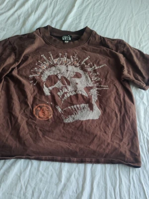 Hellstar t-shirt med dödskalletryck - Cool brun t-shirt med stort vitt dödskalletryck framtill och tryck med röda och vita detaljer på ryggen. T-shirten har rund hals och korta ärmar, perfekt för dig som gillar edgy streetstyle.