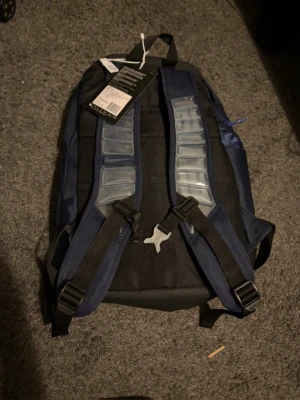 Mörkblå ryggsäck med reflexdetaljer - Snygg mörkblå ryggsäck med stora fack och reflexdetaljer på framsidan. Justerbara axelband med vaddering och smart bröstspänne. Materialet är slitstark textil och botten är förstärkt. Perfekt för skola eller träning.