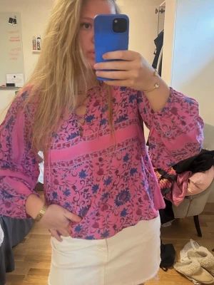 Rosa blommig blus med lång ärm - Säljer en rosa blus med blått blommönster och dekorativa band vid halsen. Blusen har avslappnad passform, rund halsringning och långa ärmar med resår vid handlederna. Perfekt för dig som gillar färg och mönster.