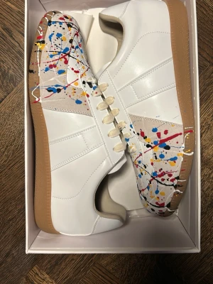 Vita sneakers med färgstänk - Unika vita sneakers med beige detaljer och färgstänk i rött, blått, gult och svart. Skorna har vita snören, gummisula i brunt och en klassisk låg siluett. Perfekta för dig som vill sticka ut med en kreativ och modern stil.