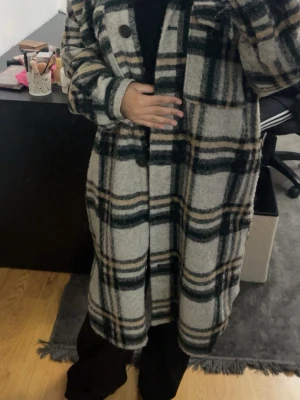 Lång rutig ullkappa i grått och grönt - Snygg lång kappa med rutigt mönster i grått, grönt och beige. Kappan har stora knappar framtill, bröstficka och en avslappnad passform. Perfekt för kyliga dagar och ger en trendig look med sitt oversized snitt och mjuka ullblandning.