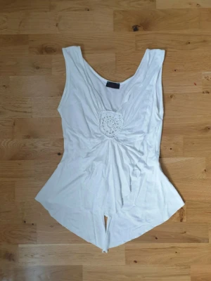 Vit ärmlös topp med broderi - Säljer en vit ärmlös topp med v-ringning och spetsdetalj framtill. Toppen har en asymmetrisk nederkant som ger en unik look och är tillverkad i ett mjukt material. Perfekt för dig som vill ha något enkelt men ändå speciellt.  Y2k