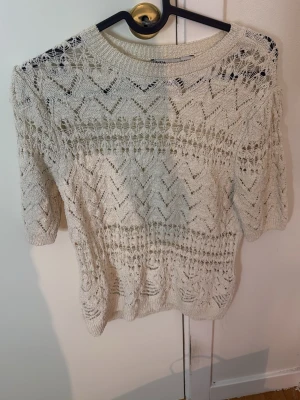 Vit stickad topp från ZARA - Säljer en vit stickad topp från ZARA med korta ärmar och rund hals. Tröjan har ett genombrutet, spetsliknande mönster i zigzag och är tillverkad i ett mjukt, tunt material. Perfekt för lager på lager och en trendig look.
