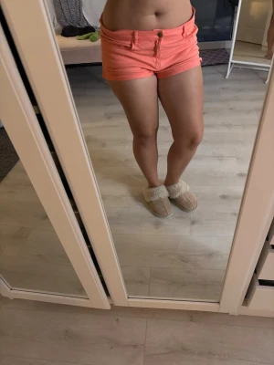 Korta korallfärgade shorts - Säljer ett par snygga, korta shorts i en fräsch korallfärg. De har låg midja, klassisk femficksdesign och stängs med knapp och dragkedja framtill. Perfekta för varma sommardagar och ger en trendig look till din outfit.