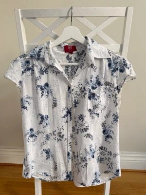 Vit blommig kortärmad skjorta - Snygg vit skjorta med blått blommigt mönster och korta ärmar. Skjortan har klassisk krage, bröstficka och knappar framtill. Perfekt för en fräsch och somrig look. Skriv vid frågor 🩵
