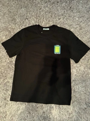 Svart t-shirt med grafiskt tryck - Svart t-shirt med färgstarkt grafiskt tryck och texten 'Casa Blanca' på både bröstet och ryggen. T-shirten har rund hals och korta ärmar, och är tillverkad i mjuk bomull. Perfekt för en trendig och avslappnad stil. 