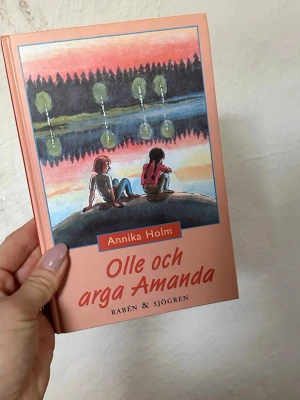 Olle och arga Amanda bok - En ungdomsroman med hård pärm i ljusrosa ton, skriven av Annika Holm och utgiven av Rabén & Sjögren. Omslaget har en illustrerad bild av två personer vid en sjö i solnedgång. Perfekt för dig som gillar berättelser om vänskap och känslor.