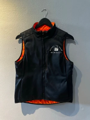 Svart dunväst med orange foder Nike - Snygg svart dunväst från Nike med orange foder och tryck på bröstet med texten 'Ready to Roar Bengals'. Västen har hög krage, dragkedja framtill och quiltad baksida. Perfekt för lager på lager och streetwear-stil.