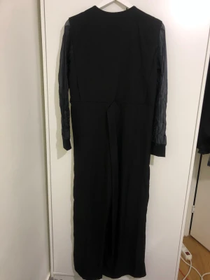 Svart långklänning med transparenta ärmar - Snygg svart långklänning med transparenta, lätt mönstrade långärmar och bred krage. Klänningen har knäppning framtill och markerad midja. Perfekt för dig som vill ha en stilren och unik look.