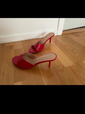 Röda slip-in pumps med smal klack strl 41 - Oanvända Snygga röda slip-in pumps med öppen tå och smal klack. Skorna har en enkel och stilren design med bred rem över foten och en feminin siluett. Materialet ser ut att vara konstläder med en blank finish. Perfekta för dig som vill sticka ut med färg.