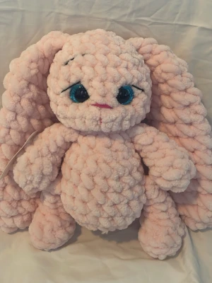 Mysig virkad rosa kanin m långa öron - Supermjuk och fluffig kanin i ljusrosa plysch med stora blå glittriga ögon och långa öron. Kaninen har en rund kropp och sött broderat ansikte. Perfekt som kramkompis eller dekoration i rummet. 23 cm