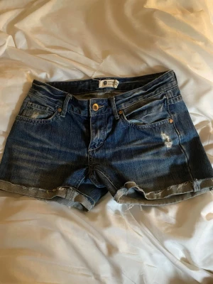 Blå jeansshorts med slitningar - Säljer ett par klassiska blå lowwaist jeansshorts i storlek 25/32 med råa kanter och slitna detaljer.