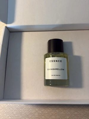 ESSNCE Carashmellow  - Parfymflaska från ESSNCE med doften Carashmellow. Flaskan är cylindrisk i genomskinligt glas med svart lock och vit etikett med svart text. Modern och stilren design som passar perfekt på parfymhyllan.