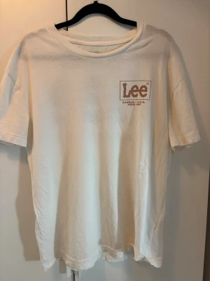 Vit t-shirt från Lee med tryck - Vit t-shirt från Lee med brunt tryck på bröstet och stort Lee-tryck på ryggen. Klassisk rund hals och korta ärmar. Skön och enkel modell i bomull, perfekt till jeans eller shorts.
