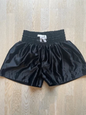 Svarta boxningsshorts med resår - Snygga svarta boxningsshorts i glansigt material med bred resår i midjan och en vit logga framtill. Perfekta för träning eller match. Klassisk passform och korta ben ger bra rörelsefrihet.