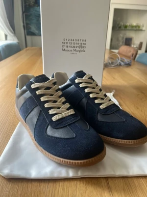 Maison Margiela blå sneakers - Snygga sneakers från Maison Margiela i marinblå mocka och läder med beige snören och klassisk gummisula. Ikonisk design med stilrena detaljer och låg profil. Perfekta för dig som vill ha något unikt och trendigt i garderoben. (DUSTBAG KOMMER INTE MED)