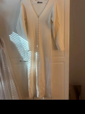 Vit stickad långklänning med knappar - Elegant vit långklänning med v-ringning och dekorativa knappar längs hela framsidan. Klänningen har trekvartsärm och är tillverkad i ett mjukt tyg som ger en skön känsla. Perfekt för sommarens alla tillfällen.