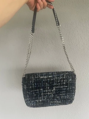 Mörkblå tweed axelväska med kedja - Snygg axelväska i mörkblå tweed med inslag av vitt och ljusblått. Väskan har en silverfärgad kedjerem med svart axelparti i läderimitation. Klassisk rektangulär form och lockstängning. Perfekt för dig som vill ha en trendig och stilren accessoar.