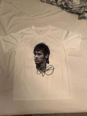 Vit t-shirt med porträtt och tryck - Vit t-shirt med svartvitt porträtt och signatur på framsidan. Baksidan har ett stort tryck med sköld och texten 'The prince who never became a king'. Klassisk passform och rund hals. Perfekt för dig som gillar unika prints.