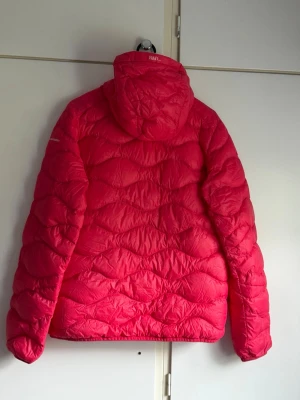 Röd dunjacka Peak Performance dam XL - Snygg röd dunjacka från Peak Performance i dammodell, storlek XL. Jackan har huva och quiltad design som ger en sportig look. Perfekt för kalla vinterdagar och håller dig varm med sitt fluffiga dunfoder.