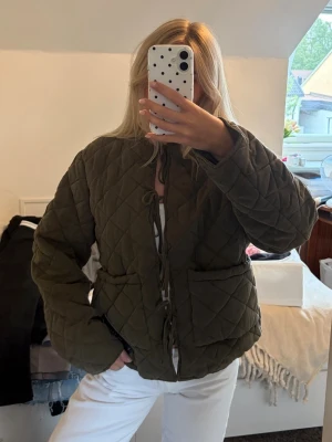 Olivgrön quiltad jacka med snörning - Quiltad jacka i olivgrönt med snörning framtill och två stora fickor. Jackan har en loose passform och långa ärmar. Materialet känns mjukt och ger en trendig vibe till din outfit. Orginalpris är 1099kr. 