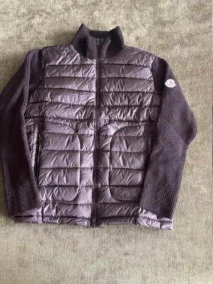 Svart cardigan  moncler - Snygg grå cardiganjacka med quiltad, vadderad framsida och stickade ärmar samt rygg. Jackan har hög krage, två stora fickor framtill och dragkedja. Perfekt lager-på-lager-plagg med modern look och Moncler-logga på ärmen.