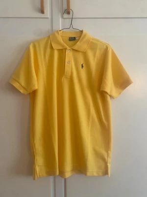 Polo Ralph Lauren var pikétröja,  - Snygg Polo Ralph Lauren gul pikétröja med klassisk krage och korta ärmar. Tröjan har knappar framtill och en liten broderad logga på bröstet. Perfekt för en fräsch och sportig look.
