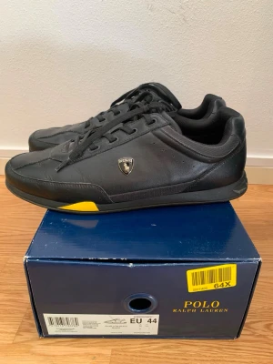 Svarta sneakers Polo Ralph Lauren - Stilrena svarta sneakers från Polo Ralph Lauren med diskret gul detalj på sulan och Polo-logga på sidan. Skorna har lågt skaft, snörning och är tillverkade i slätt lädermaterial. Perfekta för en clean och sportig look.