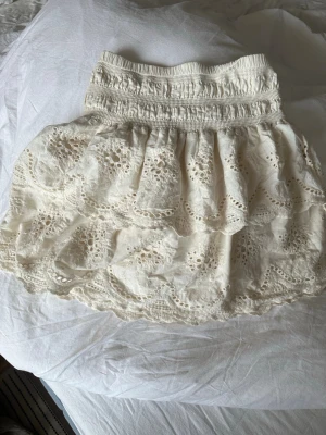 Cremé Vit volangkjol med spetsdetaljer - Söt kort créme vit kjol med bred resår i midjan och flera lager volanger. Kjolen har fina spetsdetaljer och broderade hålmönster som ger en romantisk känsla. Perfekt för dig som gillar en feminin och somrig stil.