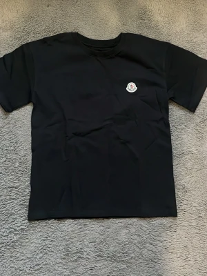 Svart t-shirt med liten logga - En stilren svart t-shirt med rund hals och korta ärmar. På bröstet sitter en liten broderad logga som ger en clean och exklusiv känsla. Perfekt till jeans eller shorts för en enkel men snygg look.
