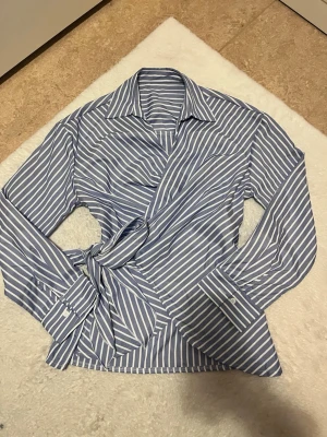 Blåvit omlottblus med knytband - Snygg blå och vit randig omlottblus med långa ärmar och klassisk krage. Blusen har knytband i midjan och breda manschetter med knappar. Perfekt för en trendig och stilren look.
