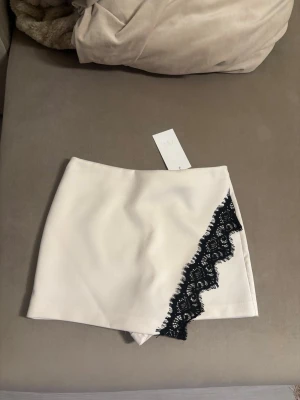Virala byxkjol Zara  - Supersnygg vit skort från Zara med unik svart spetsdetalj längs ena sidan framtill. Skorten har en stilren, rak passform och dragkedja bak. Perfekt för dig som vill ha kjol-looken men med shortskomfort. Materialet är slätt och känns lyxigt.