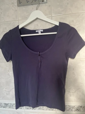 Mörklila kortärmad topp med knappar - En enkel och 2000s inspirerad topp från Subdued med en super fin mörklila färg. Toppen har korta ärmar och två knappar framtill samt en rundad urringning och är gjord i mjukt och bekvämt material. Perfekt bas topp som passar till mycket!💕