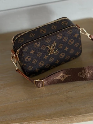 Brun monogram handväska med bred rem - Snygg handväska i mörkbrunt konstläder med guldiga monogram och detaljer. Väskan har två dragkedjefack, en ytterficka och en bred, justerbar axelrem i brunt tyg med matchande mönster. Perfekt för dig som vill ha en trendig accessoar med plats för det viktigaste.