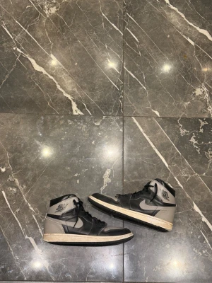 Jordan 1 high shadow  - Snygga höga sneakers i svart och grått med klassisk siluett och snörning. Skorna har en tydlig swoosh på sidan och detaljer vid ankeln. Yttermaterialet ser ut att vara en mix av läder och syntet. Perfekta för dig som gillar streetwear och vill sticka ut.