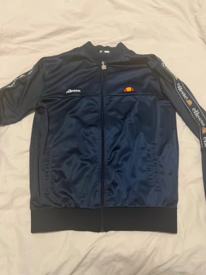 Mörkblå ziptröja från Ellesse - Snygg mörkblå ziptröja från Ellesse med logga på bröstet och vita Ellesse-detaljer längs ärmarna. Tröjan har hög krage, hel dragkedja och ribbade muddar. Tillverkad i glansigt material som ger en sportig retrostil.