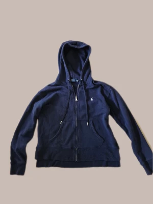 Mörkblå hoodie från Polo Ralph Lauren - Snygg mörkblå hoodie från Polo Ralph Lauren med dragkedja framtill, två fickor och klassisk huva med dragsko. Liten broderad logga på bröstet. Tillverkad i mjukt bomullsmaterial som känns skönt mot huden. Perfekt för en avslappnad och stilren look.