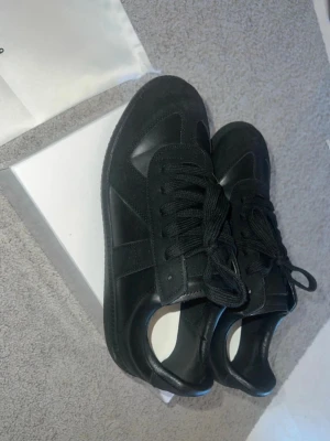 Svarta sneakers från Maison Margiela - Stilrena svarta sneakers från Maison Margiela i en mix av mocka och läder. Klassisk låg modell med svarta skosnören och diskreta detaljer. Perfekta för dig som gillar minimalistisk och clean stil.