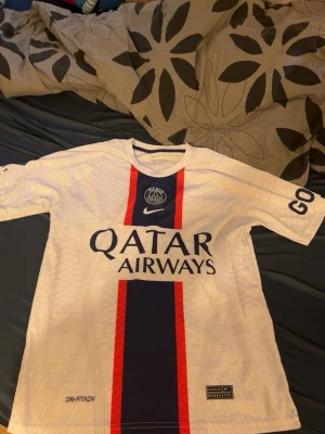 PSG Mbappé bortatröja vit - Säljer en vit PSG bortatröja med Mbappé och nummer 7 på ryggen. Tröjan har blå och röda detaljer, Qatar Airways-tryck på bröstet och Dri-FIT material för bästa komfort. Perfekt för dig som älskar fotboll och vill stötta PSG.