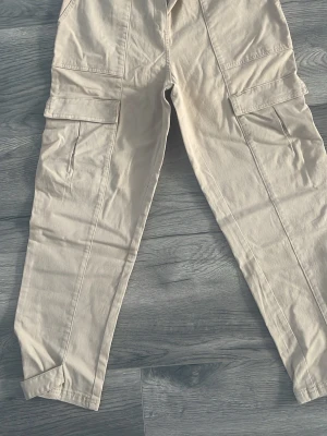 Beige cargobyxor med fickor - Snygga beige cargobyxor med raka ben och stora sidofickor på båda benen. Tillverkade i ett mjukt bomullstyg som ger en avslappnad look. Perfekta för en streetwear-stil och passar till det mesta i garderoben. Midjan 40, storlek 40