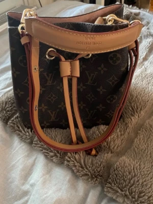 Louis Vuitton noe noe - Snygg brun väska från Louis Vuitton med klassiskt monogrammönster och detaljer i beige och guld. Väskan har justerbar axelrem och dragskostängning. Perfekt för dig som vill ha en ikonisk och rymlig handväska med lyxig känsla.