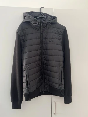 Svart hoodie-cardigan med quiltad front - Svart cardiganjacka med quiltad framsida och huva. Framsidan och huvan är vadderade medan ärmar och baksida är i mjukt stickat material. Jackan har dragkedja framtill, två fickor med dragkedja och en diskret logga på bröstet. Perfekt för lager-på-lager och streetwear. Extremt bra skick, finns inga tecken på användning.