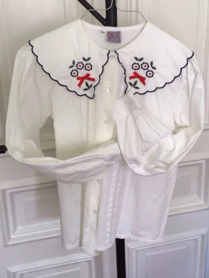 Vit skjorta med broderad krage - Vit skjorta med lång ärm och knappar framtill. Kragen har broderade blommor och röda rosetter, vilket ger en unik och söt detalj. Skjortan är i bomull och har en klassisk passform. Perfekt för dig som gillar romantiska detaljer.