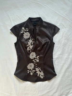 Vintage topp - Snygg topp i glansigt material med vita broderade blommor framtill. Blusen har korta ärmar, ståkrage och en diskret dragkedja i sidan. Figurnära passform och elegant asiatisk touch. Vid köp av flera varor får du rabatt💛