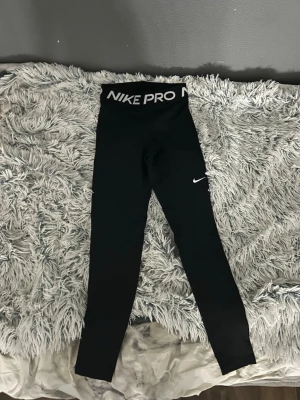 Svarta Nike Pro leggings - Svarta Nike Pro leggings med hög midja och bred resår med logga. Tight passform och stretchigt material som passar perfekt till träning. Klassisk Nike Swoosh på benet. Snygga och sportiga tights för dig som gillar att röra på dig.