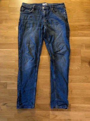 Blå raka jeans med fem fickor - Säljer ett par klassiska blå jeans med rak passform och fem fickor. Jeansen har normal midja, dragkedjegylf och knapp framtill. Materialet är denim i bomull och de har snygga slitningar för en avslappnad look.