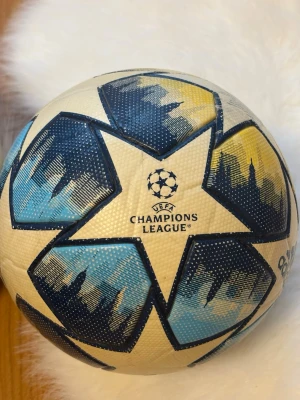 UEFA Champions League fotboll - Säljer en officiell UEFA Champions League fotboll med blå, gula och vita stjärnmönster. Bollen har tryck för Saint Petersburg 22 Final och är perfekt för matcher eller träning. Snygg design med struktur för bättre grepp och kontroll.